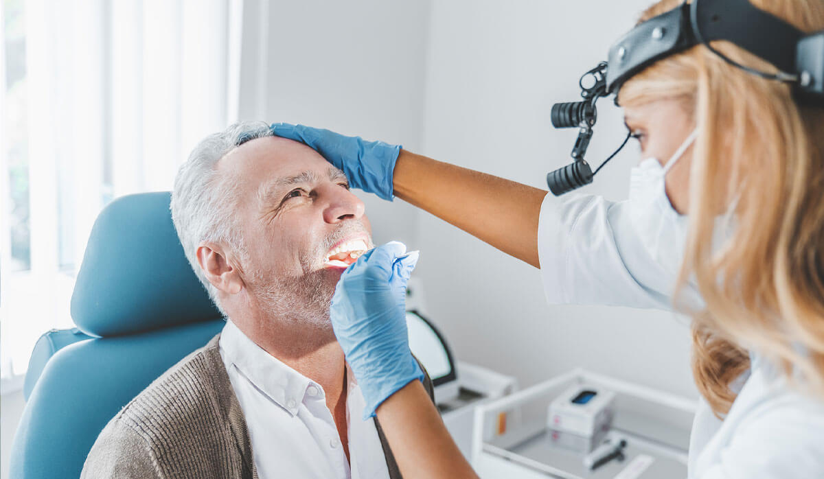 A woman dentist treating a male patient who is wearing a white shirt and brown sweater.