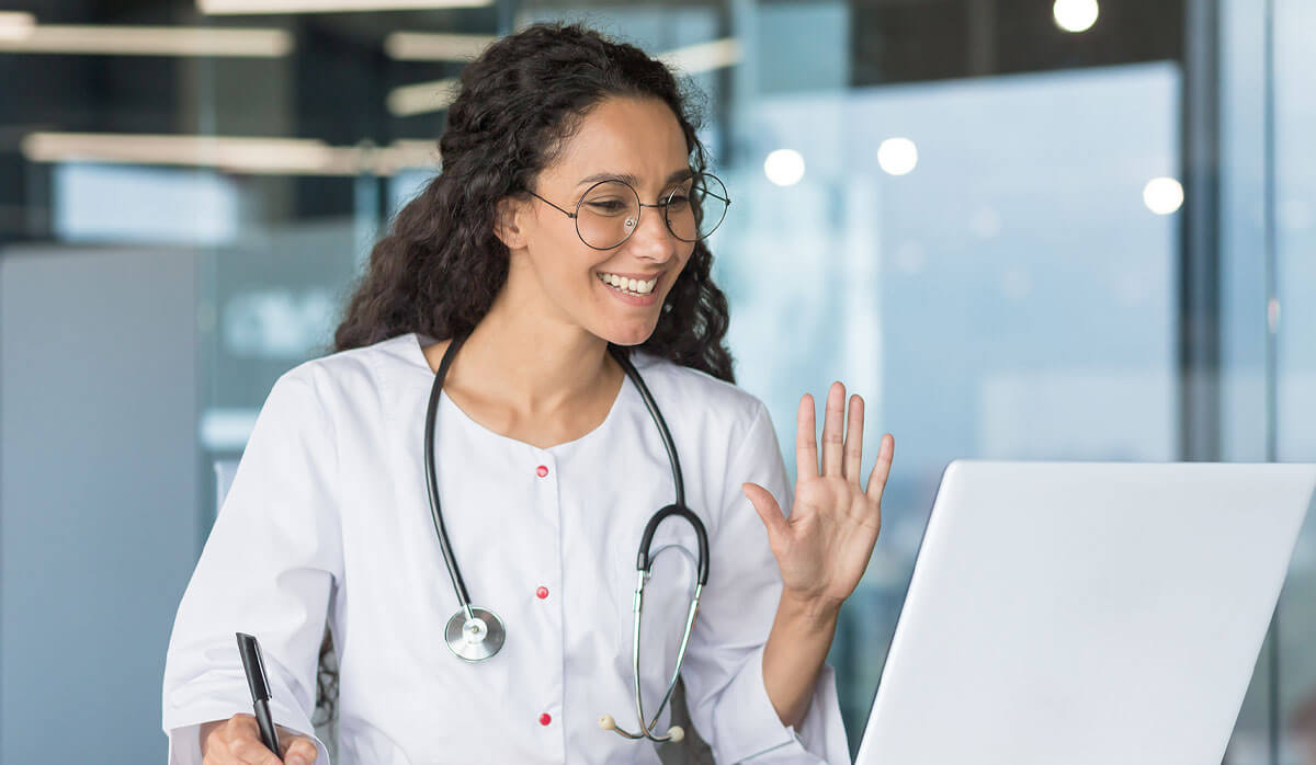 A woman with a white coat and stethoscope on her shoulders, smiling at a laptop with a pen in one hand and waiving at the screen with her other hand.